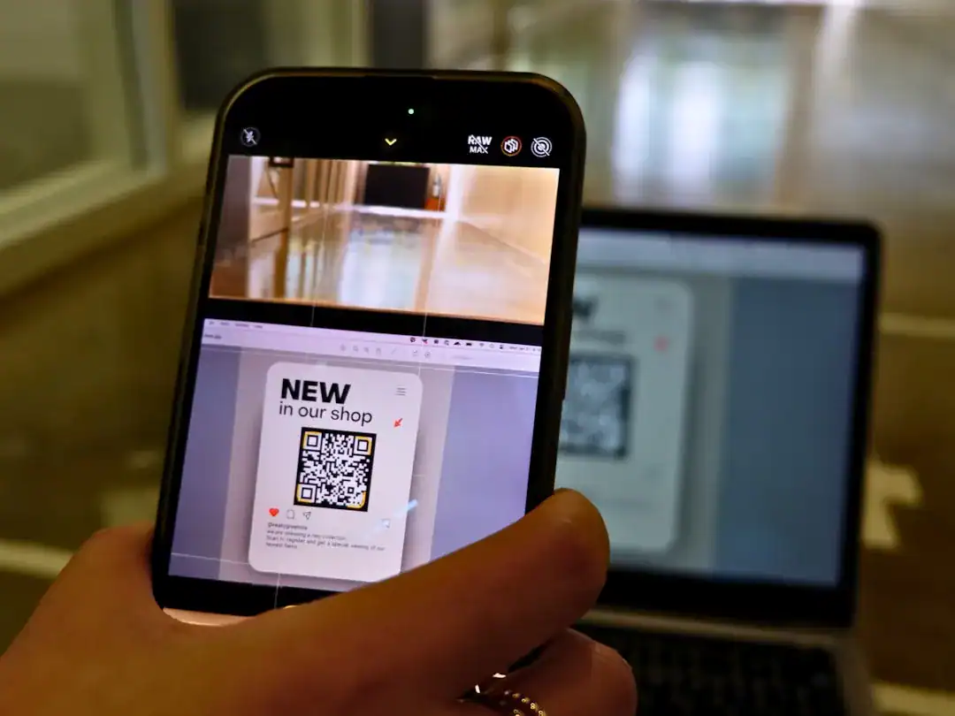 Close-up of hand holding a smartphone to scan a QR poster in a store, laptop blurred in the background