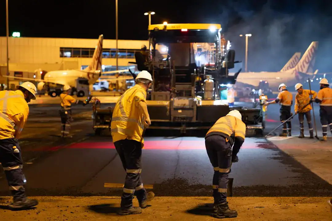 Night-time airport runway resurfacing with workers laying asphalt; planes visible in the background.