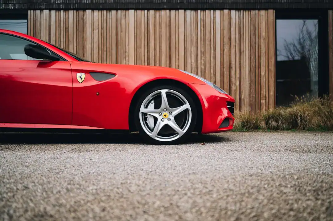 Close-up side view of a red Ferrari with alloy wheel and emblem against a wooden backdrop.