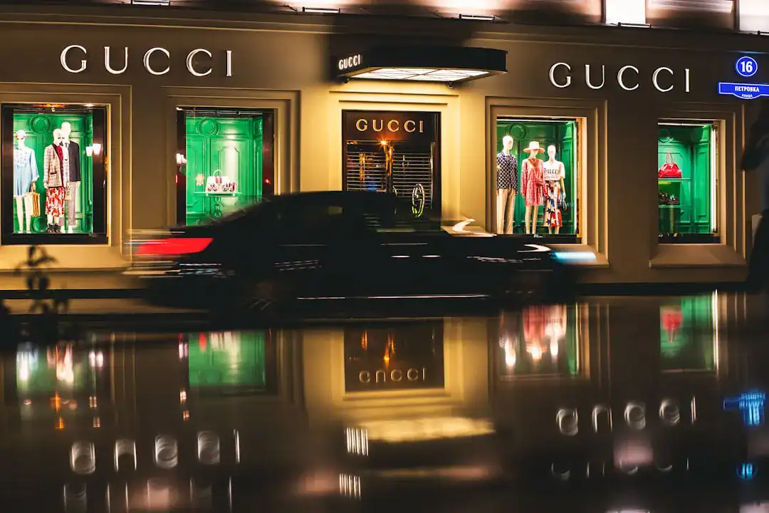 Night view of a Gucci storefront with mannequins in green-lit windows and a blurred passing car on a wet street.