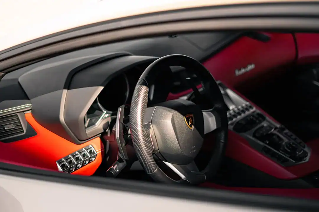 Close-up view of a Lamborghini interior with a carbon-fiber steering wheel and red-orange dashboard accents.