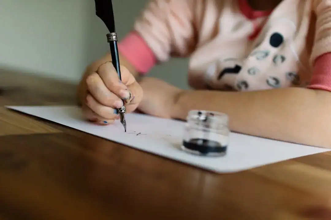 Close-up of hand writing with a dip pen and ink bottle on desk