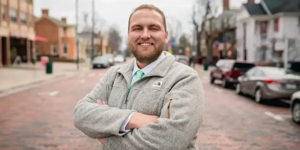 Man with arms crossed on a brick-paved street in a small town, storefronts and parked cars in the background