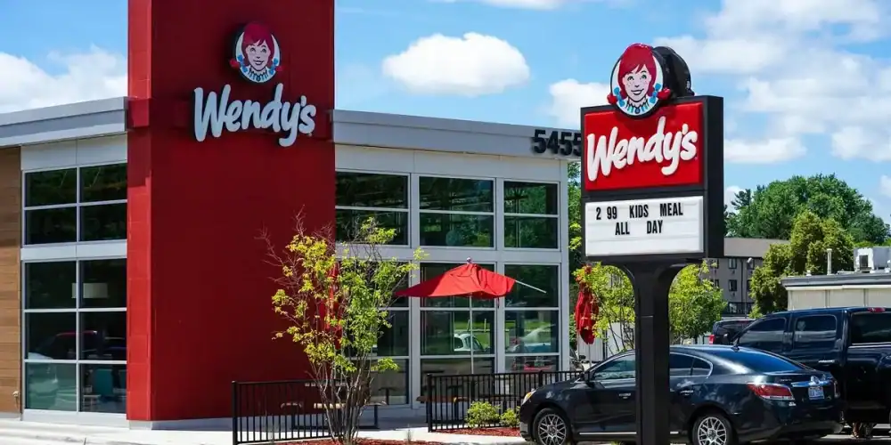 Exterior view of Wendy's with red sign tower, glass windows, outdoor seating, and parked cars