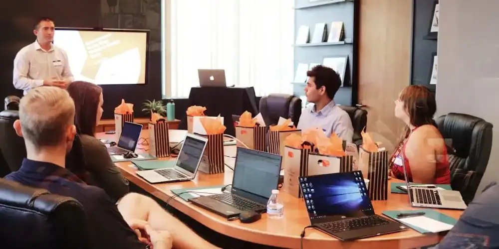 Professionals in a modern conference room sit with laptops while a presenter briefs the group beside a projection sc