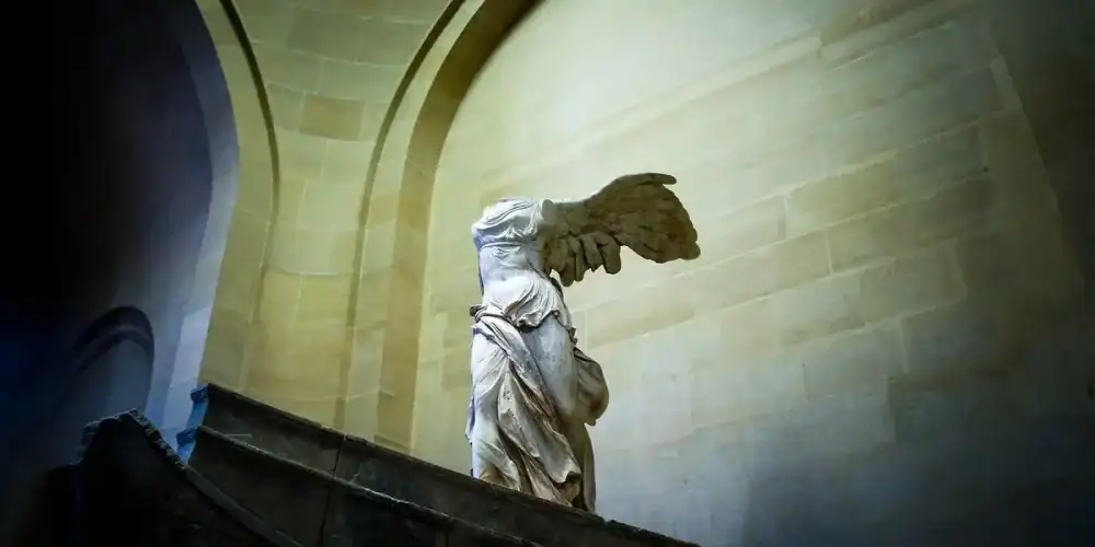 Winged statue atop a stone pedestal inside an arched interior with warm light and blue shadows