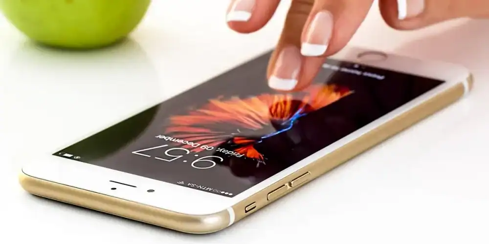 Hand with French-manicured nails tapping a gold iPhone on a white surface with a green apple in the background