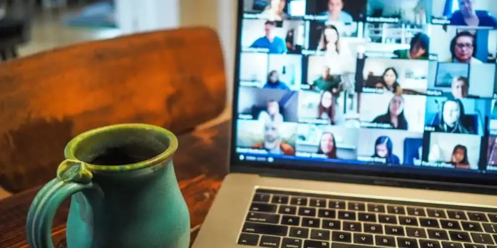 Laptop showing video conference grid and teal mug on a wooden desk