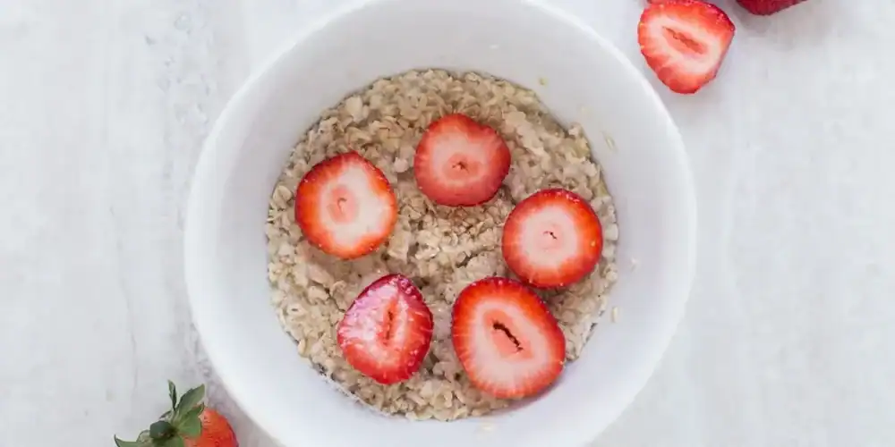 White bowl of oatmeal topped with strawberry slices on a light marble surface