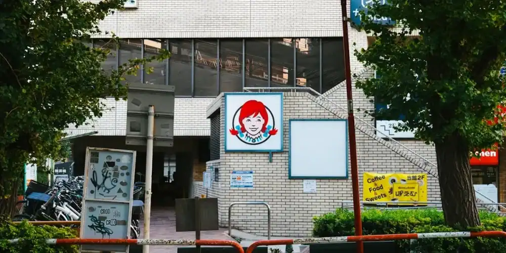 Brick storefront with a blue circular sign featuring a red-haired cartoon figure, framed by trees and a railing.