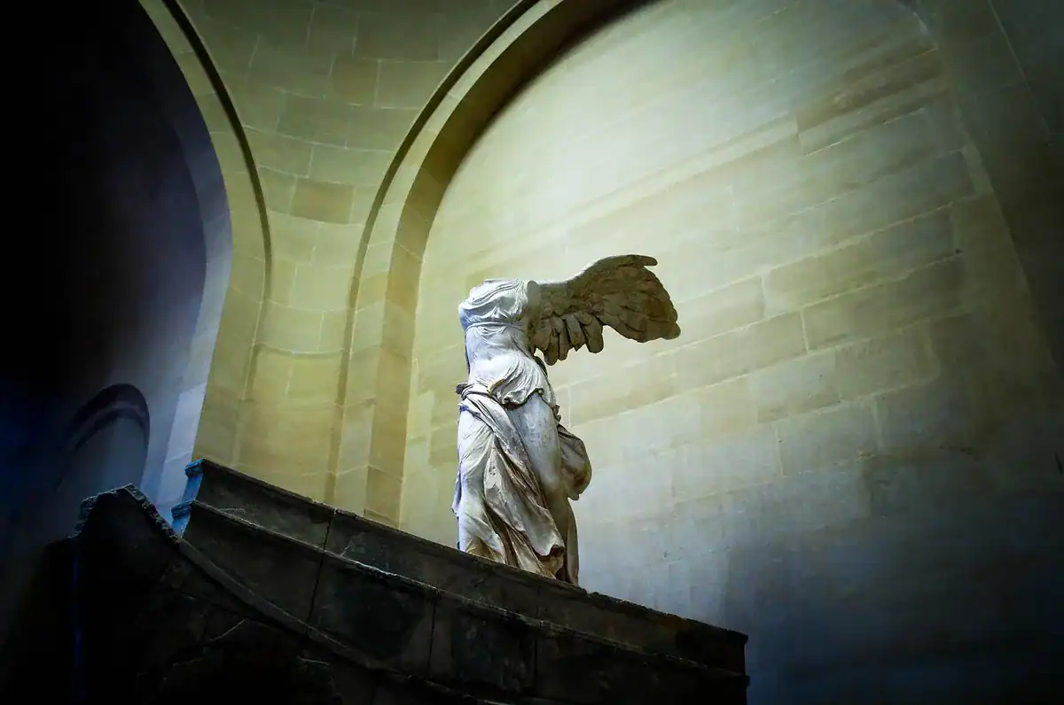 Winged statue atop a stone pedestal inside an arched interior with warm light and blue shadows