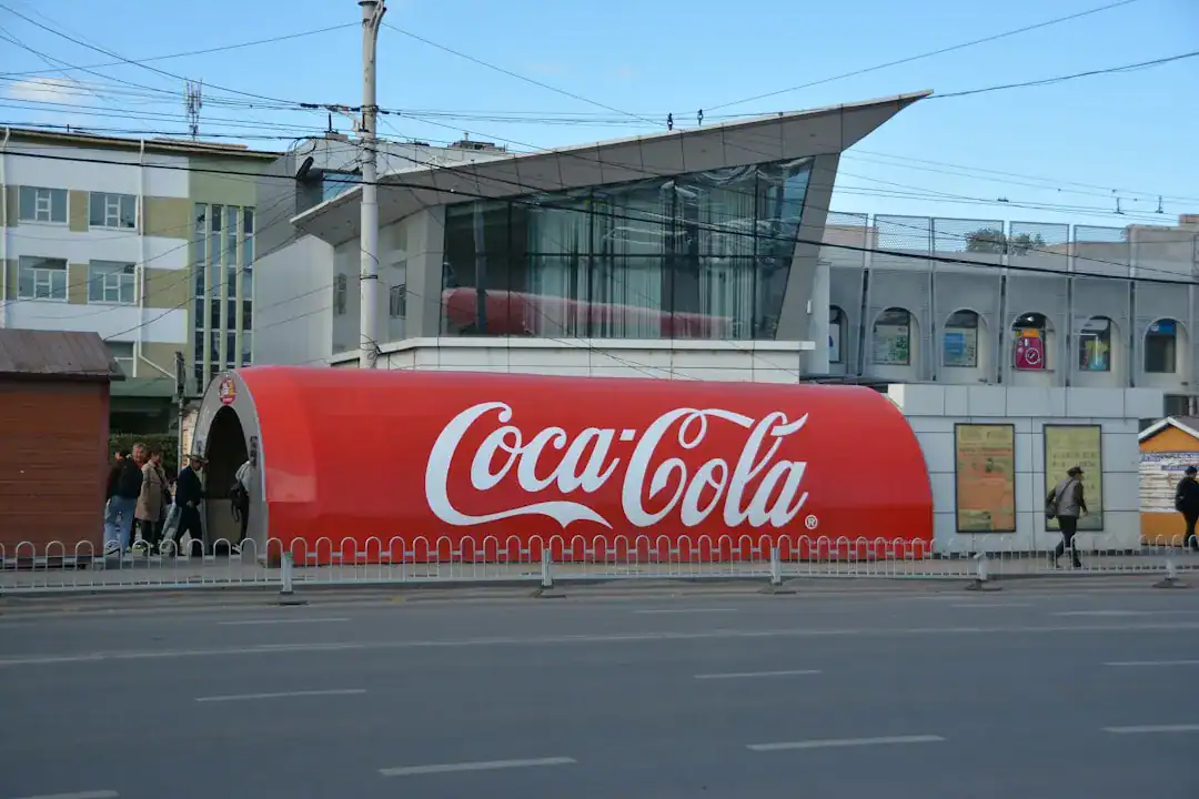 Red Coca-Cola shelter with logo on a city street, glass building behind, pedestrians nearby