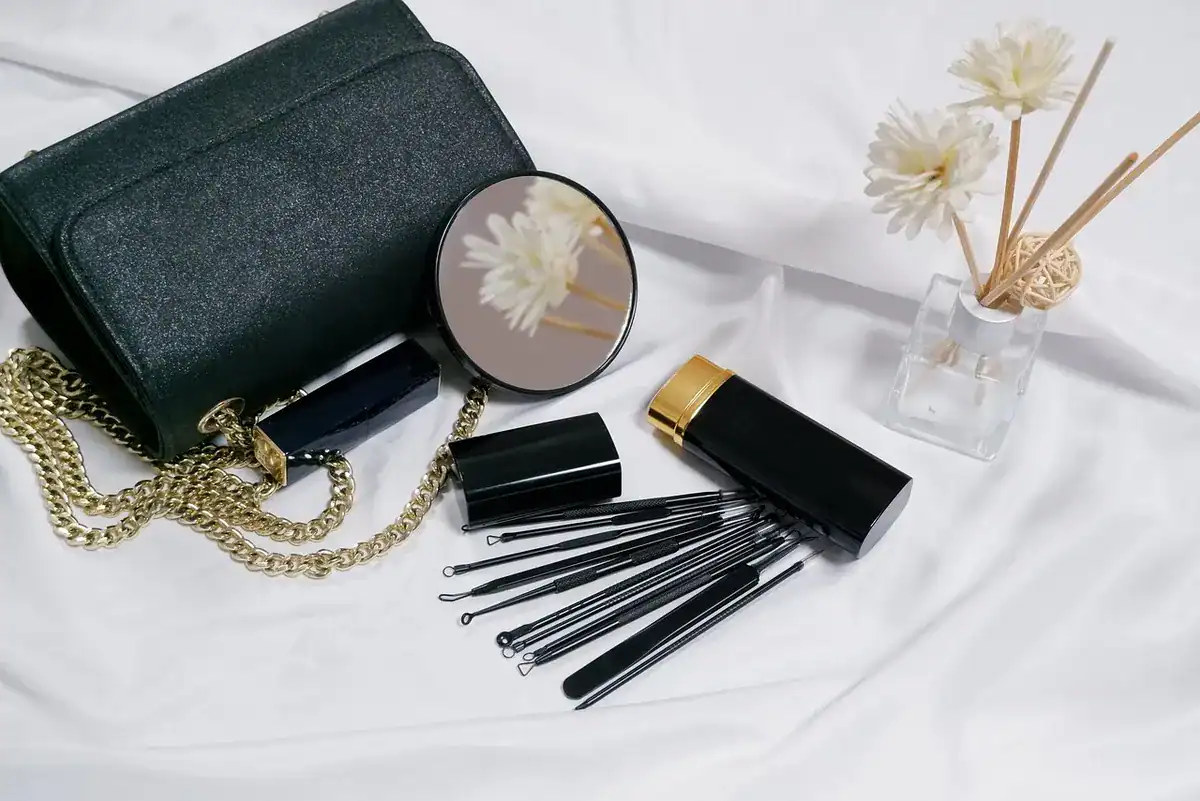 Black cosmetic bag with gold chain, makeup brushes, mirror, and diffuser sticks on white satin