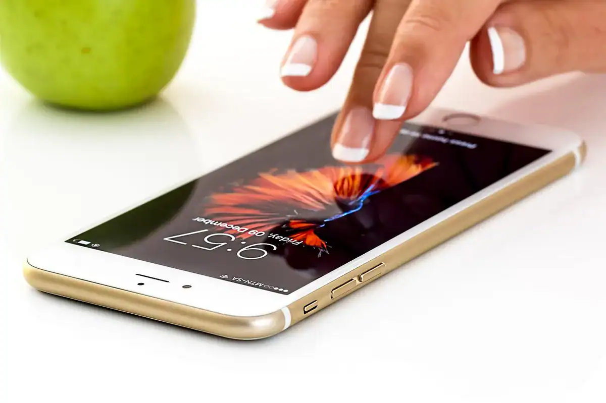 Hand with French-manicured nails tapping a gold iPhone on a white surface with a green apple in the background