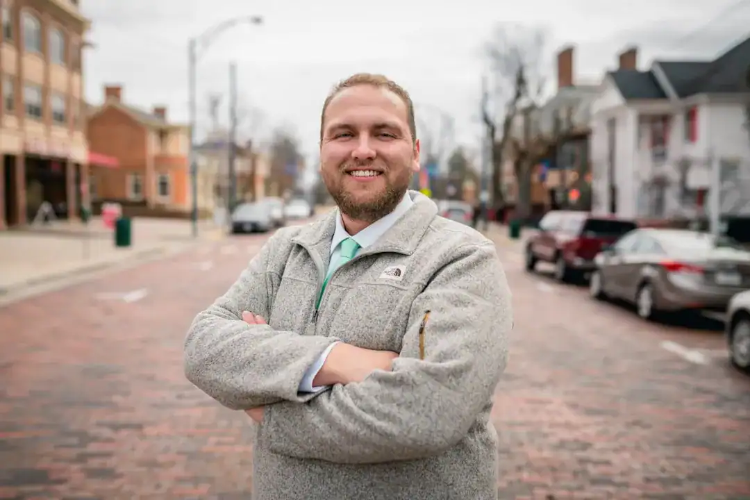 Man with arms crossed on a brick-paved street in a small town, storefronts and parked cars in the background