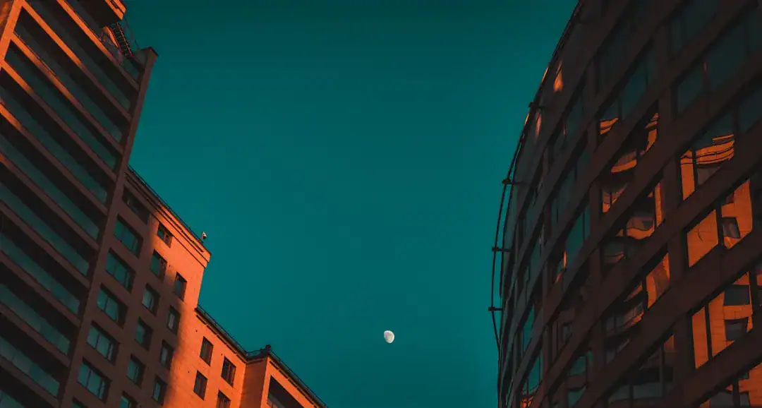 Two skyscrapers frame a teal dusk sky with a small moon between them