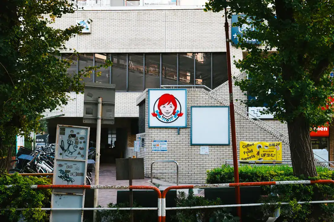 Brick storefront with a blue circular sign featuring a red-haired cartoon figure, framed by trees and a railing.