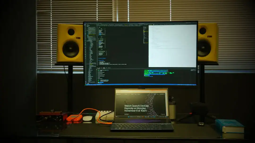 Programming desk with large monitor, laptop, and yellow studio speakers in a dim room