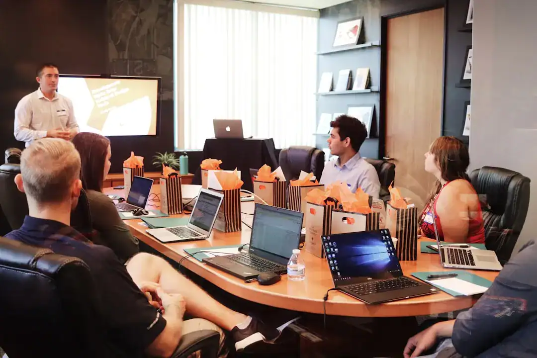 Professionals in a modern conference room sit with laptops while a presenter briefs the group beside a projection sc