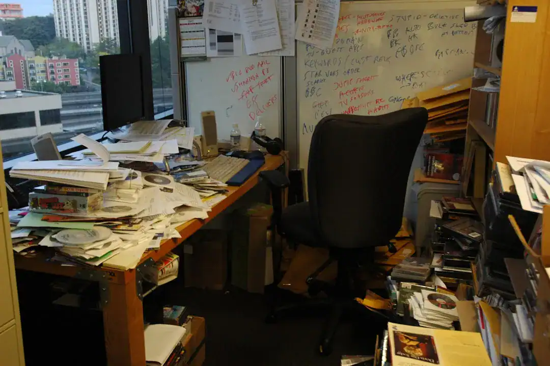Cluttered office with piles of papers, a computer, chair, and a whiteboard filled with notes.