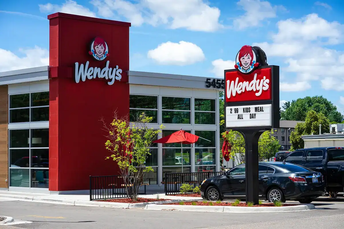 Exterior view of Wendy's with red sign tower, glass windows, outdoor seating, and parked cars