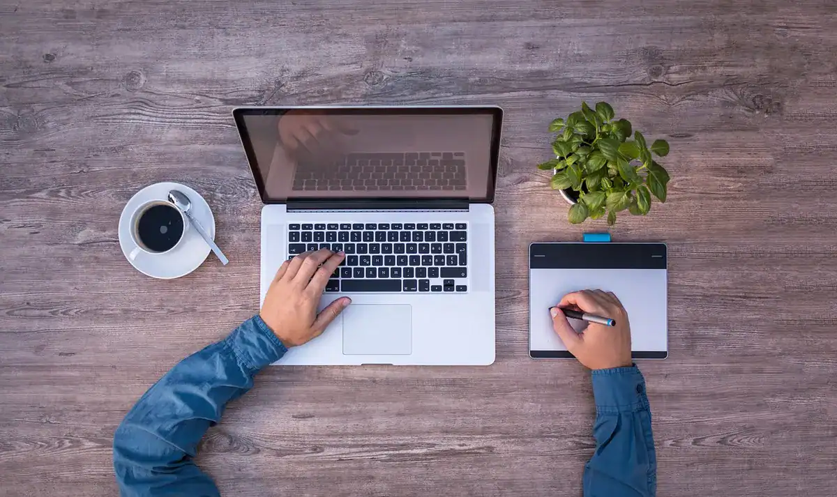 Overhead view of a laptop, drawing tablet, coffee cup, and plant on a wooden desk